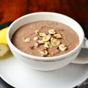 Mocha overnight oats topped with crushed hazelnuts in coffee cup on top of saucer with half banana in background.