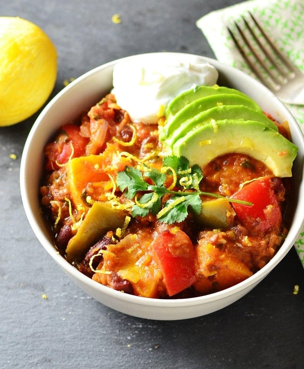 Top down view of vegetarian butternut squash chili with slices of avocado and dollop of yogurt in white bowl, with fork on top of green cloth to right and lemon to left.