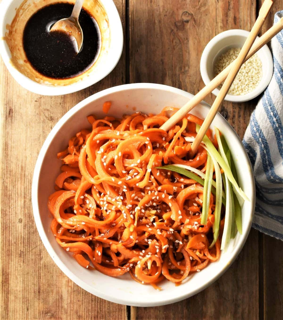 Carrot noodles stir fry in white bowl with chopsticks, seeds and glaze in background.