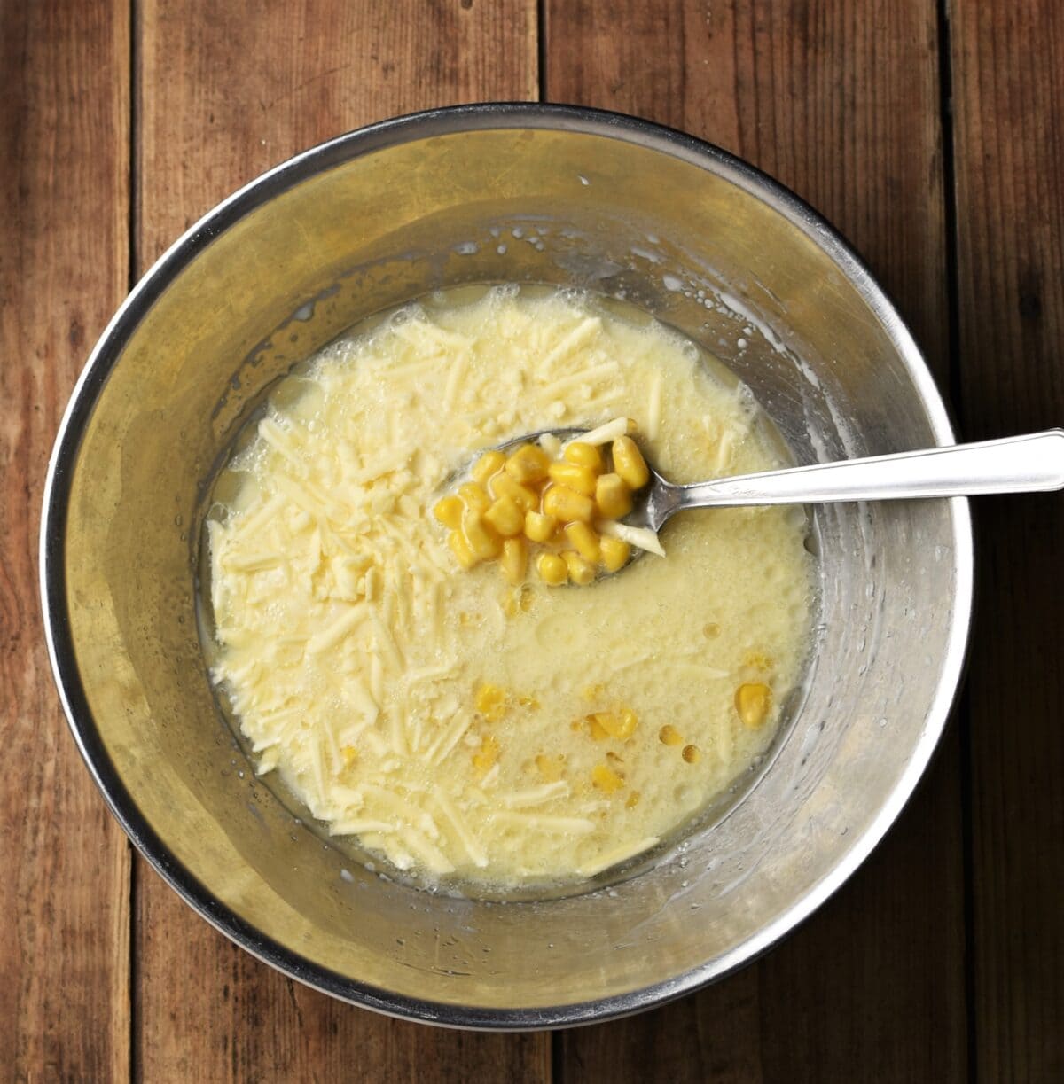 Milk mixture with sweet corn and grated cheese in large metal bowl with spoon.