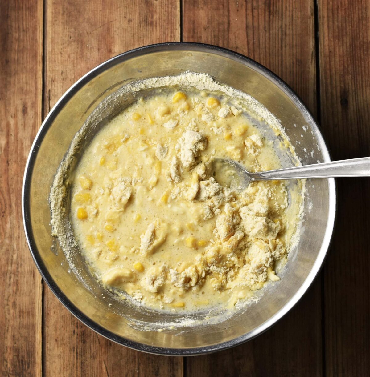 Cornmeal muffins batter in large metal bowl with spoon.