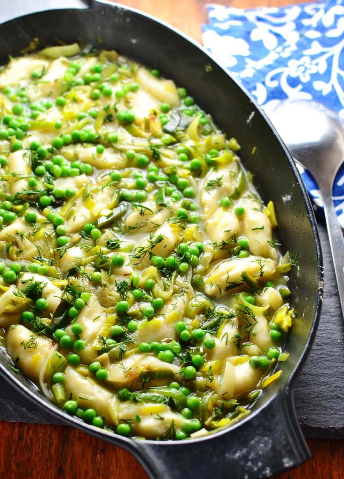 Leek stew with potato dumplings in black cast iron oval casserole dish with spoon and blue-and-white cloth in background.