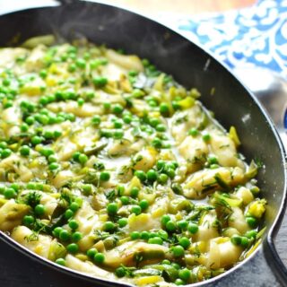 Leek stew with peas and potato dumplings in black cast iron oval casserole dish with steam visible and blue-and-white cloth in background.