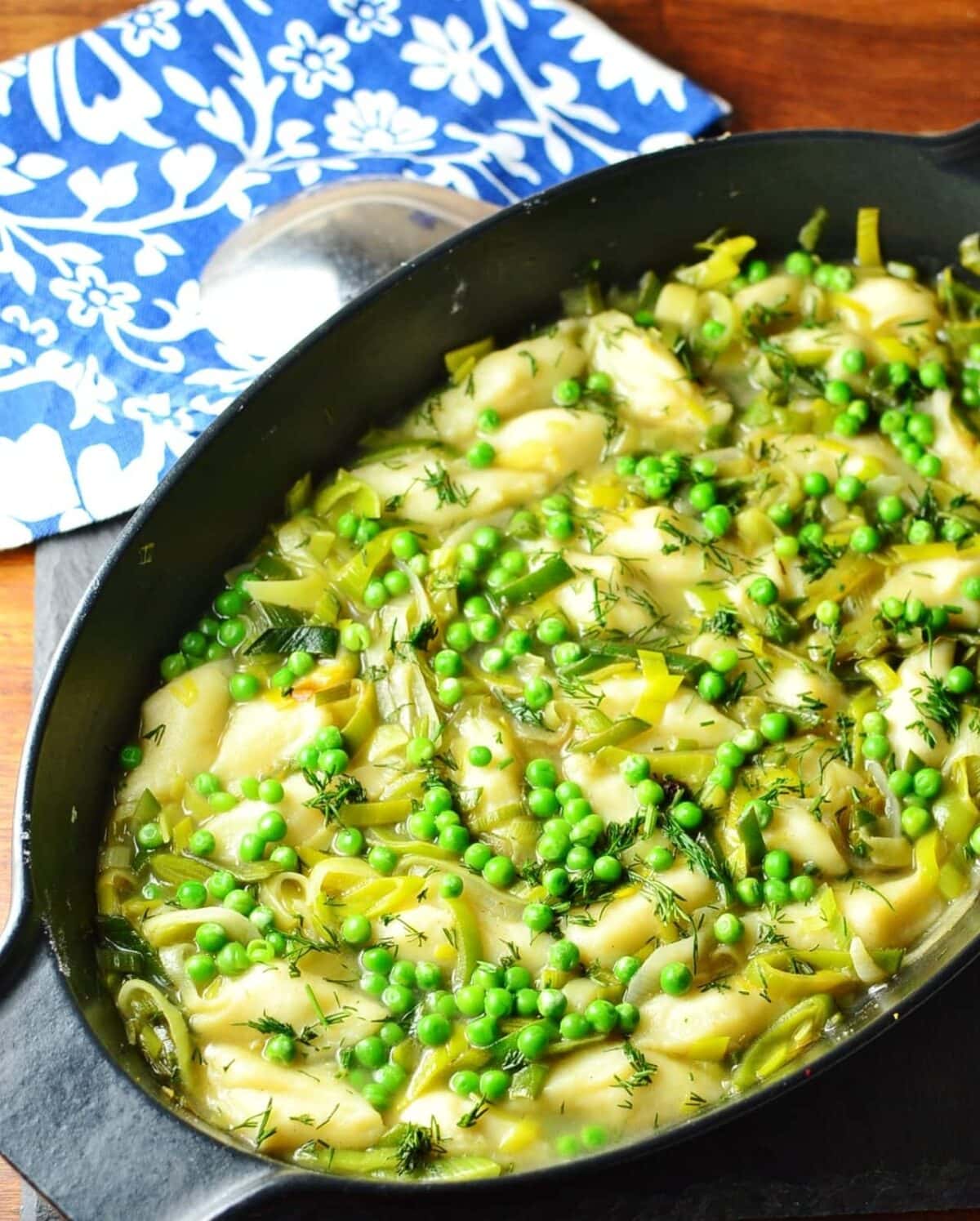 Leek stew with potato dumplings and peas in black cast iron oval casserole dish with spoon and blue-and-white cloth in background.