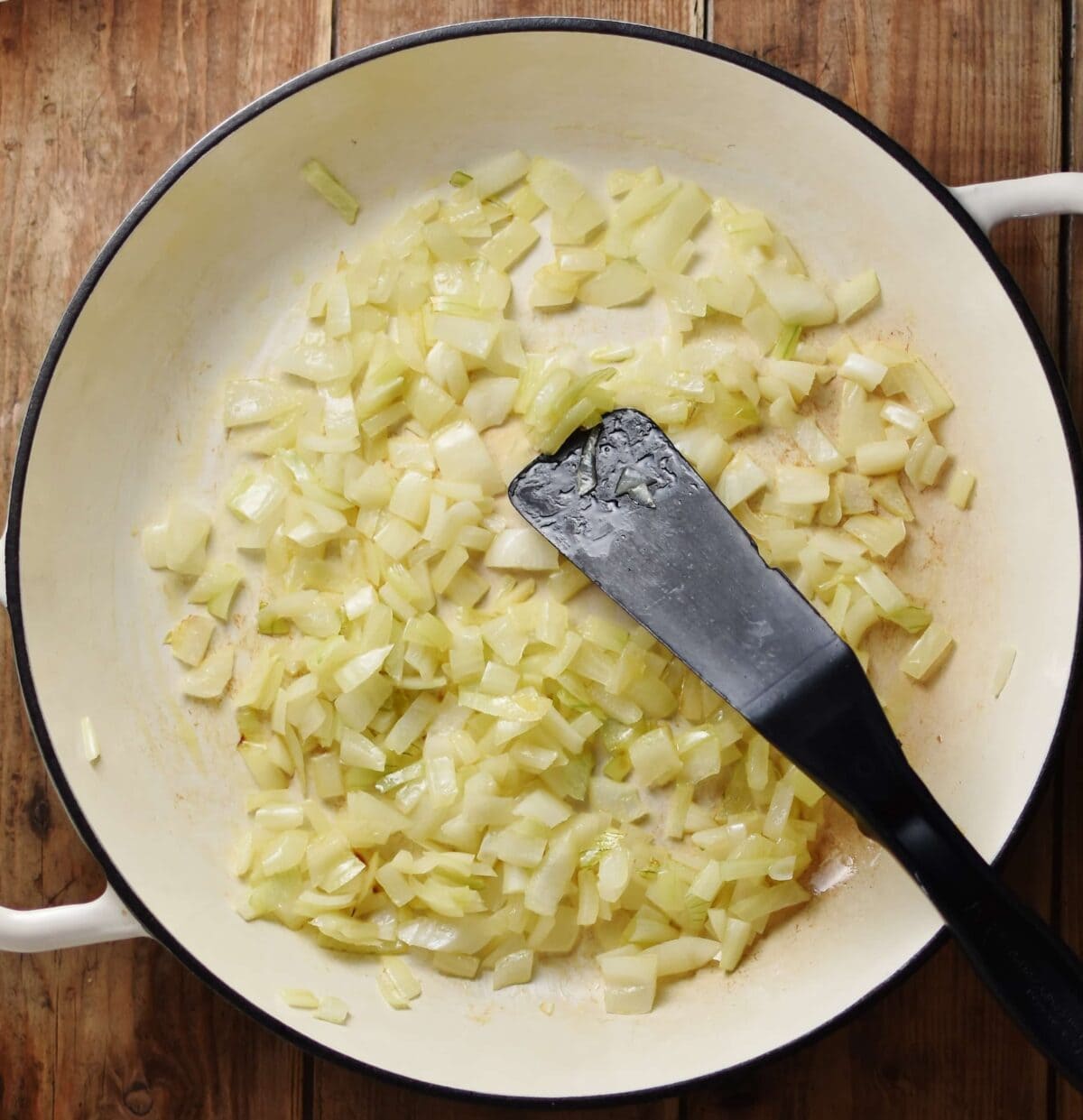 Top down view of onions frying in large shallow white pan with black spatula.