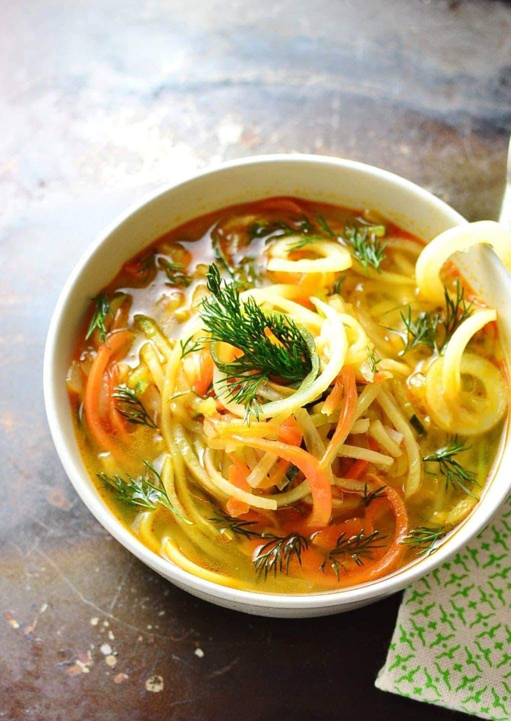 Spiralized vegetable soup in white bowl with green cloth on dark brown surface.