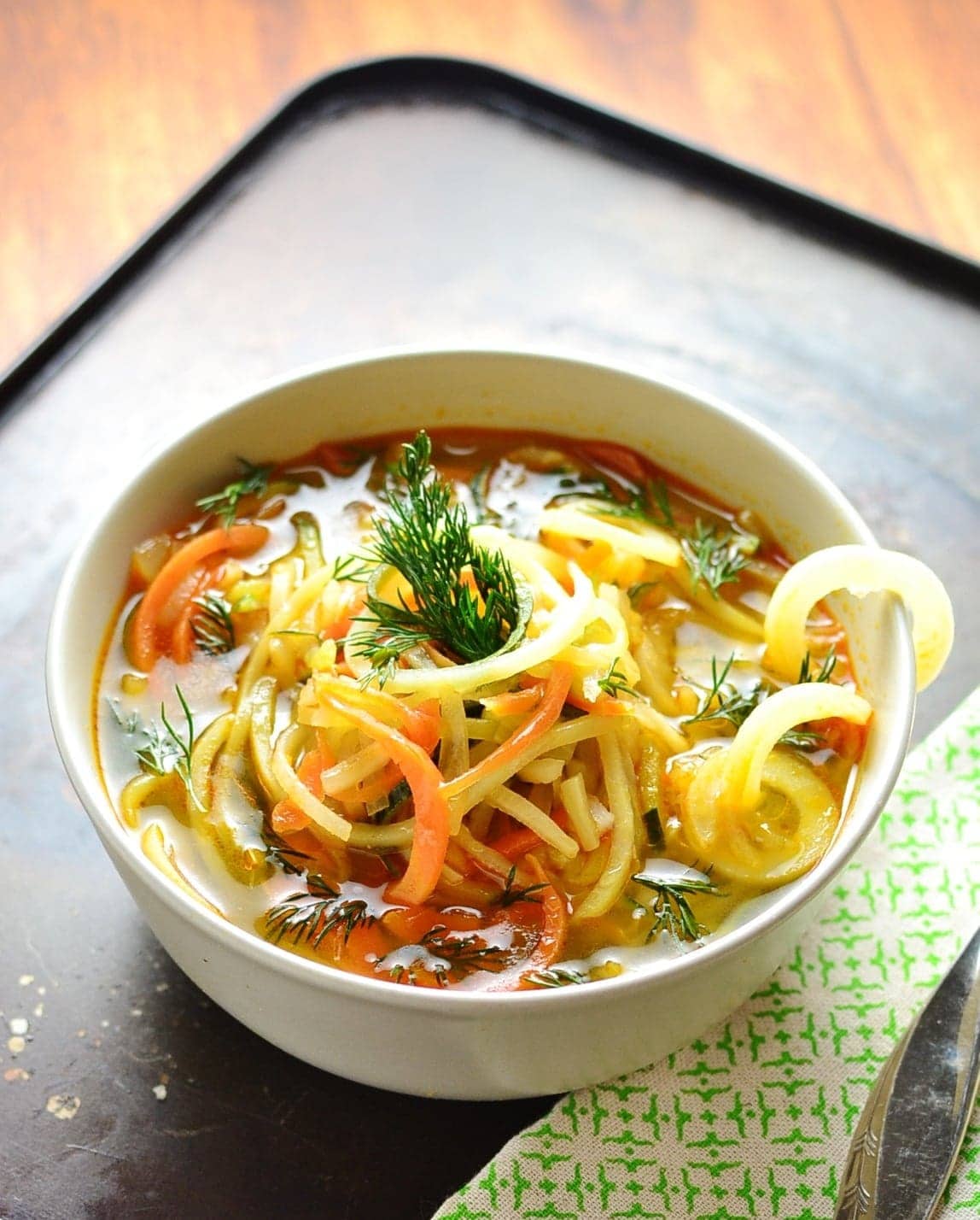 Spiralized vegetable soup in white bowl with green cloth on oven tray.