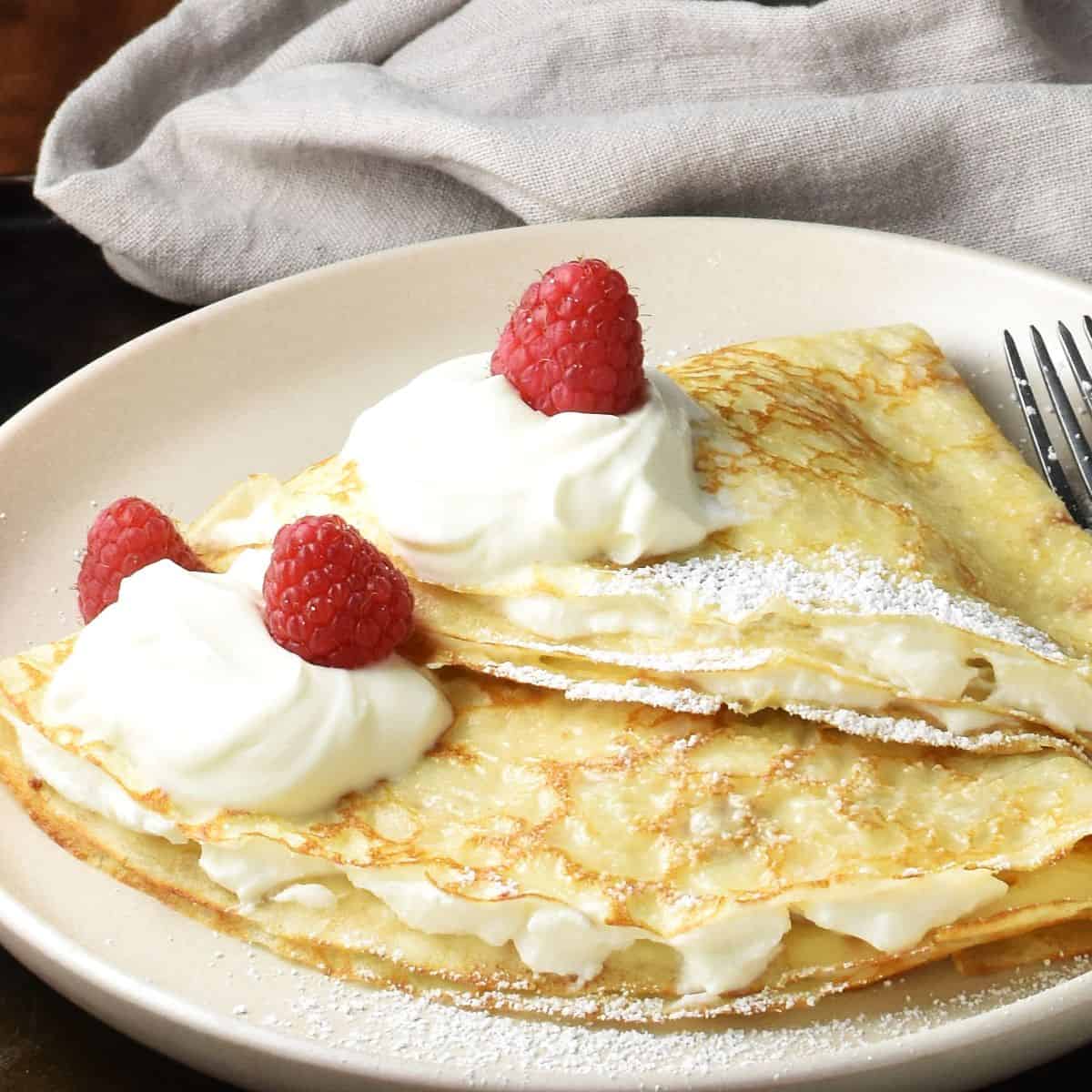 Close-up side view of 2 folded Polish nalesniki with sour cream and fruit on plate with grey cloth in background.
