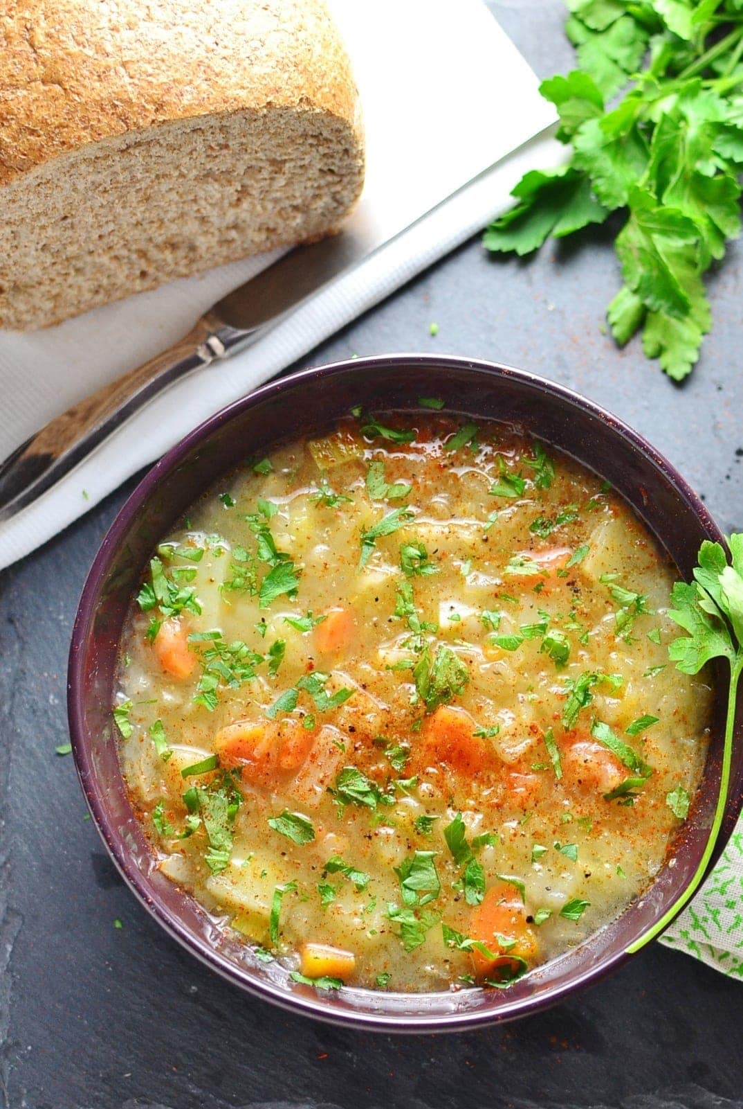 Top down view of sauerkraut vegetable kapusniak soup in purple bowl with bread loaf on white cloth with knife and parsley.
