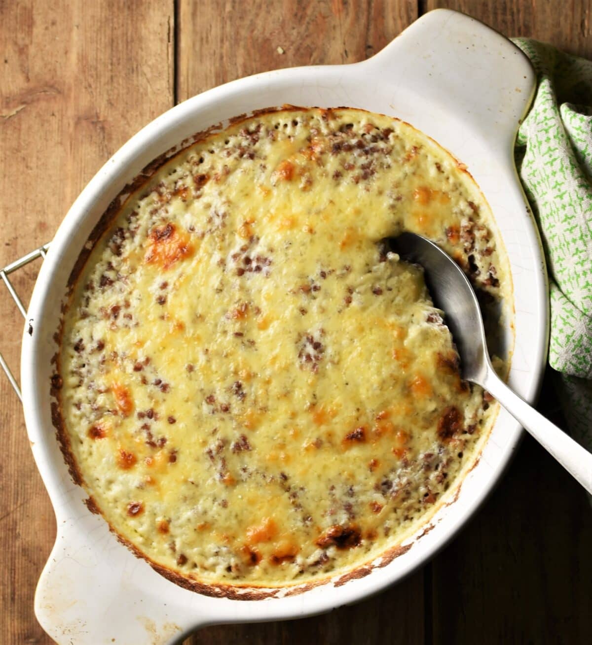 Baked buckwheat in creamy cheese sauce in white oval dish with spoon and green cloth to the right.