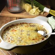 Side view of creamy buckwheat topped with melted cheese in white dish with spoon, green cloth, whisk, grated cheese and measuring cup in background.