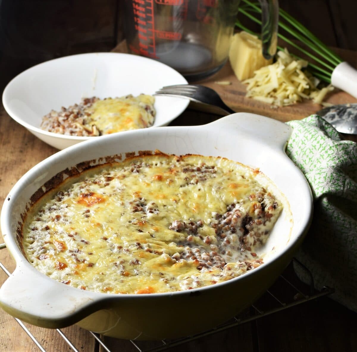 Side view of creamy buckwheat topped with melted cheese in white oval dish, white bowl with fork, grated cheese, measuring cup and whisk in background.