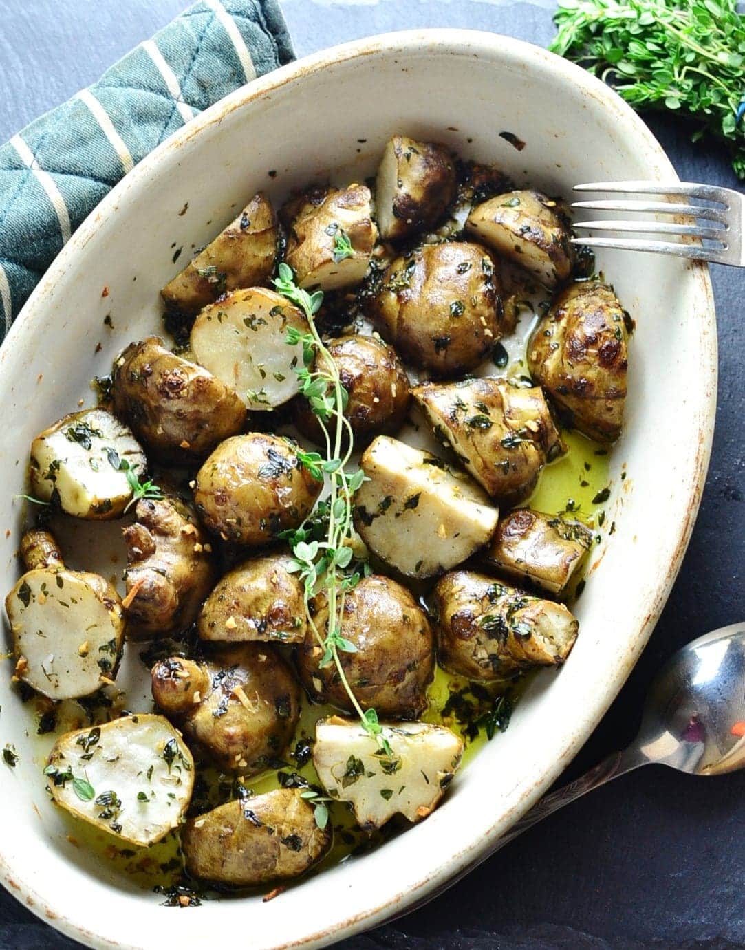 Top down view of roasted Jerusalem artichokes with herbs in white oval dish with spoon, fork and fresh herbs on dark grey table.