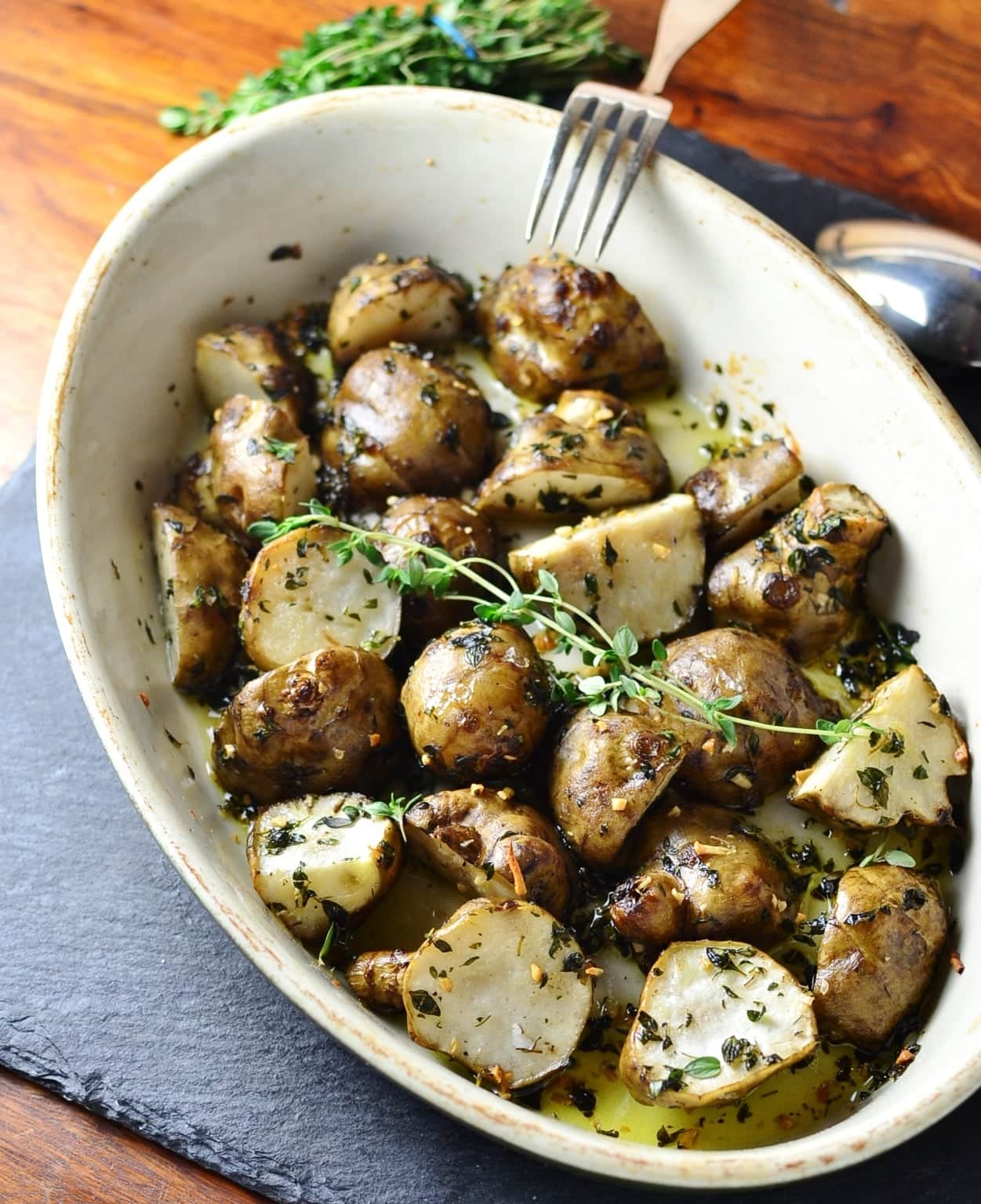 Top down view of roasted Jerusalem artichokes with herbs in white oval dish with spoon, fork and fresh herbs on dark grey table.
