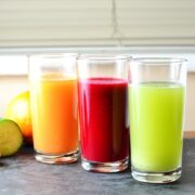 Cabbage juice in three glasses, green, red and orange, with fruit and vegetables in background on slate surface.