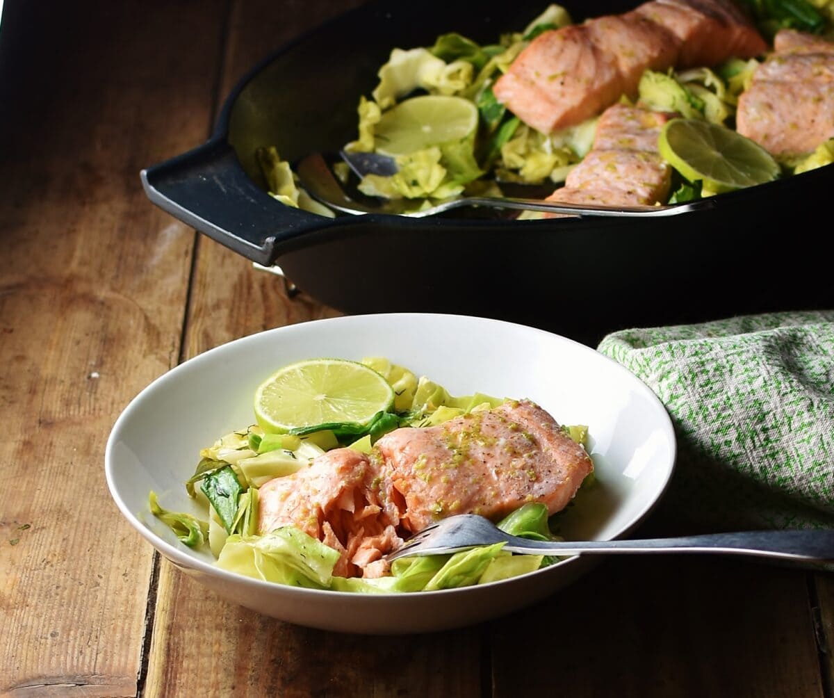 Side view of salmon and cabbage in white bowl with fork, green cloth and black dish with salmon and cabbage in background.