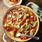 Top down view of bean, vegetable and fennel casserole with pieces of feta in large shallow white dish with spoon, green cloth in top right and casserole in small bowl at the top.