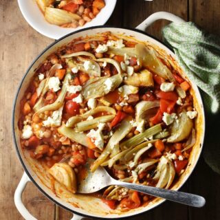 Top down view of bean, vegetable and fennel casserole with pieces of feta in large shallow white dish with spoon, green cloth in top right and casserole in small bowl at the top.