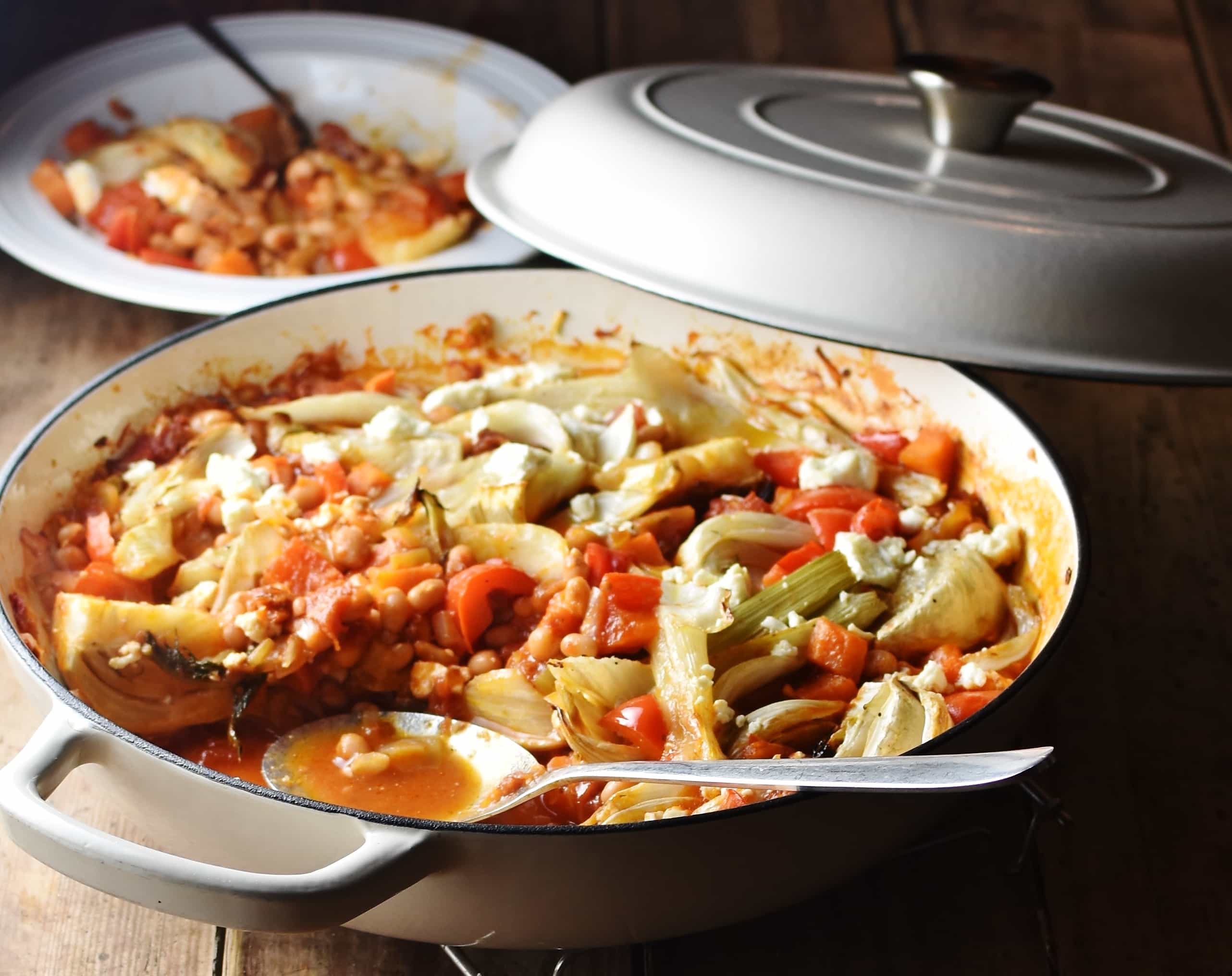 Side view of fennel vegetable bake with beans in large white shallow dish with spoon, white lid and casserole in white plate in background.