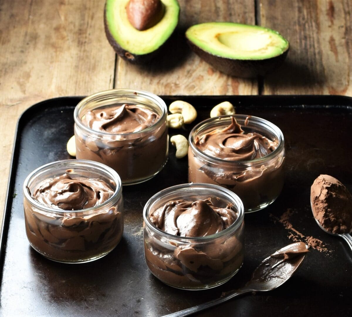 Side view of healthy chocolate mousse in 4 round dishes, 2 spoons with cocoa powder, cashews and halved avocado in background.