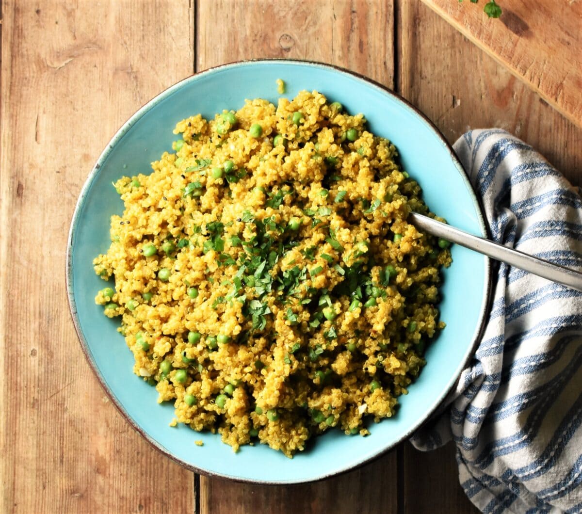 Top down view of curry quinoa with peas in blue bowl with spoon and stripy blue-and-white cloth to the right.