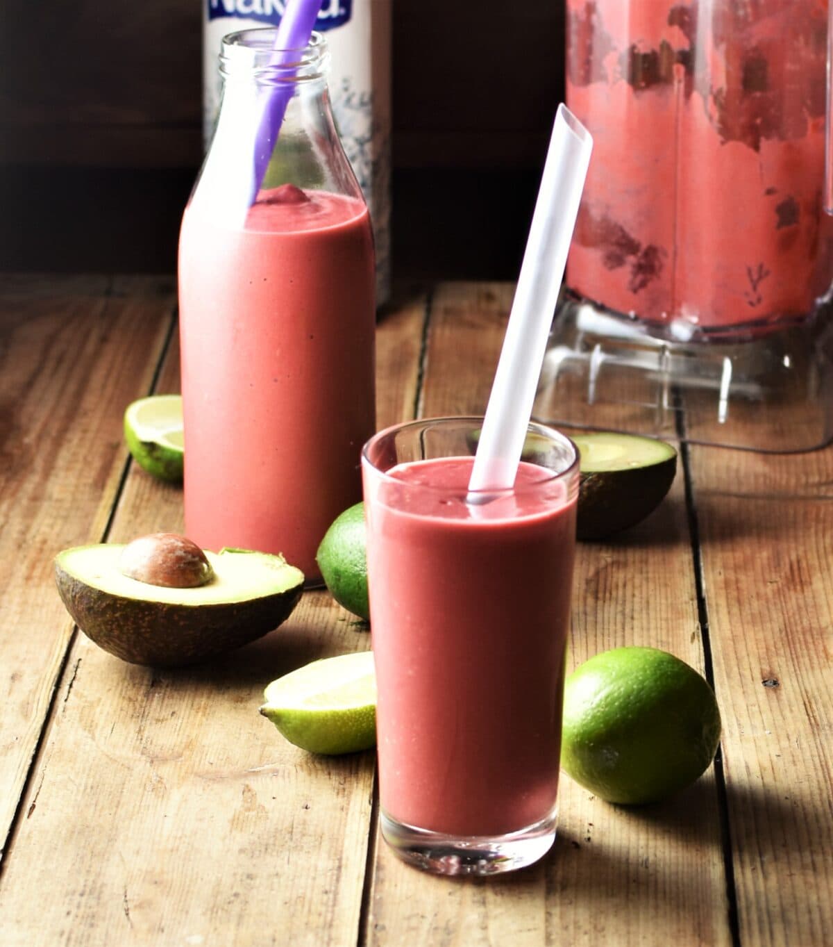 Side view of pink avocado smoothie in glass with straw, limes, avocado, smoothie in bottle and in blender in background.