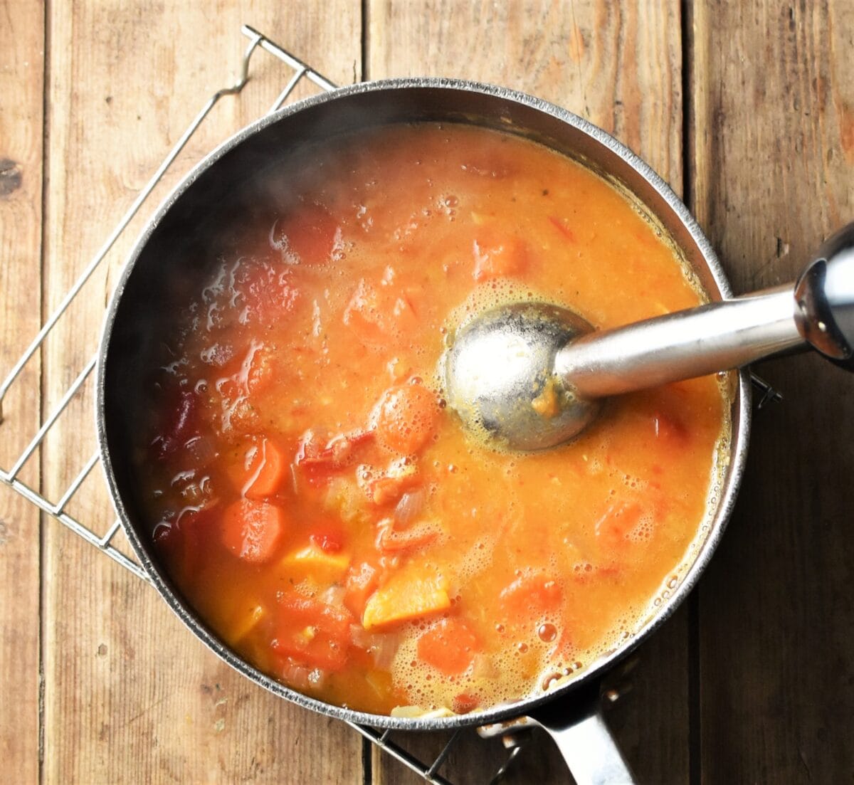 Partly pureed red vegetable soup in large pot with stick blender end piece.
