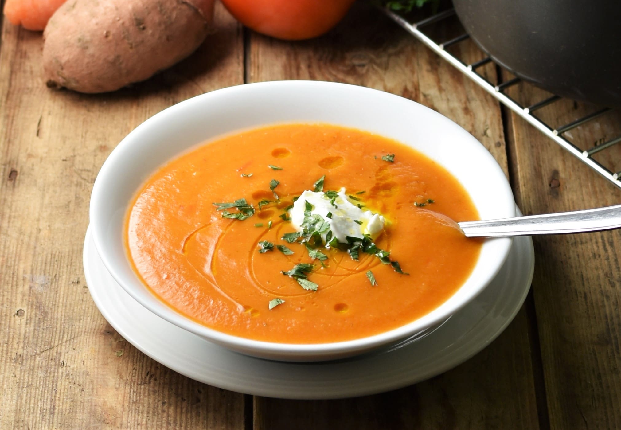 Side view of creamy red soup in white bowl with spoon on top of white plate, with vegetables in background.