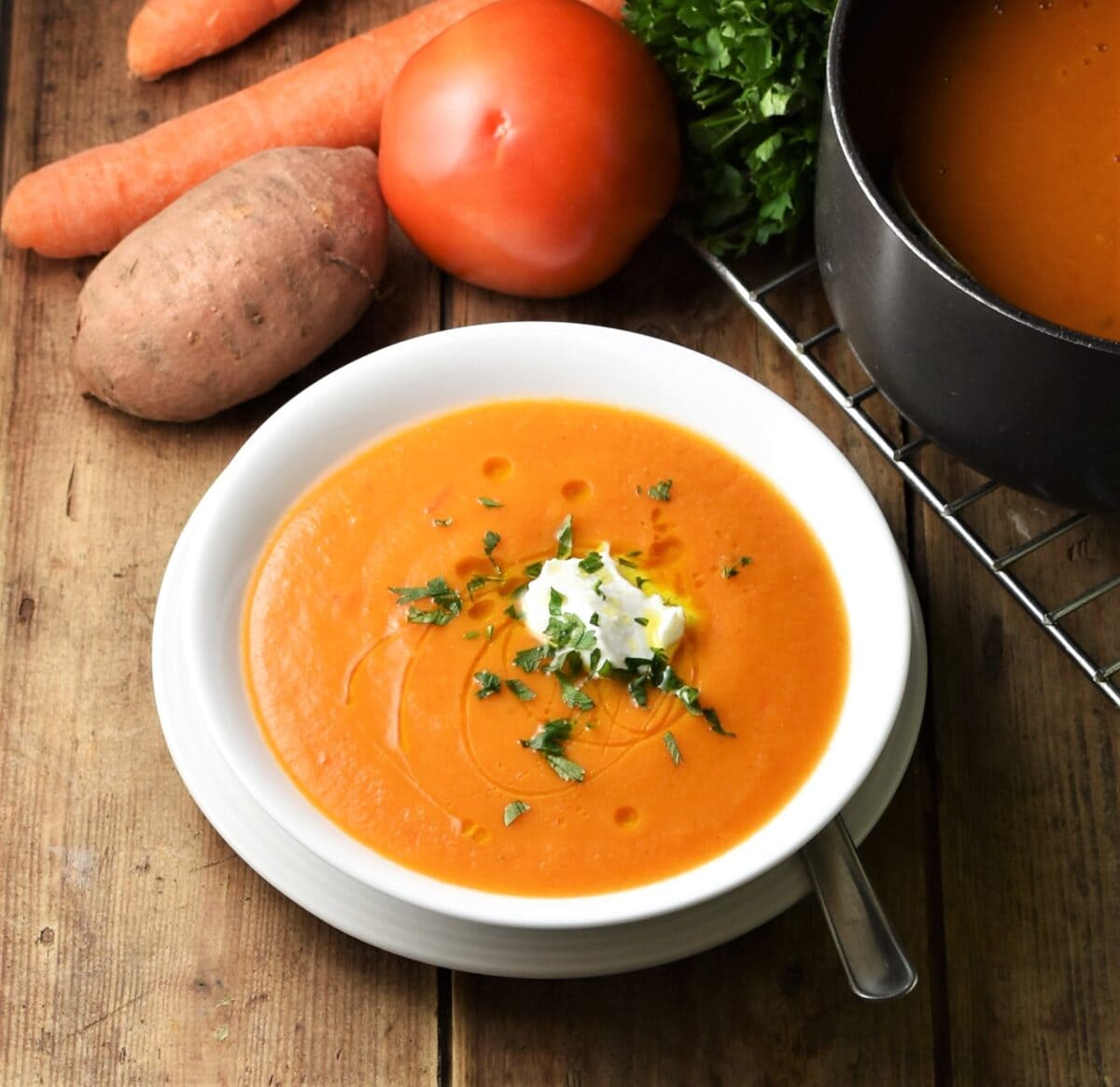 Creamy red soup in white bowl on white plate, with tomato, sweet potato, carrots and soup pot in background.