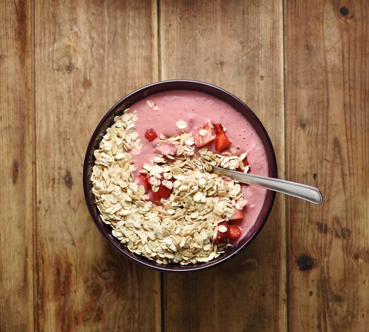 Top down view of strawberry mixture, oats and chopped strawberries with spoon in purple bowl.