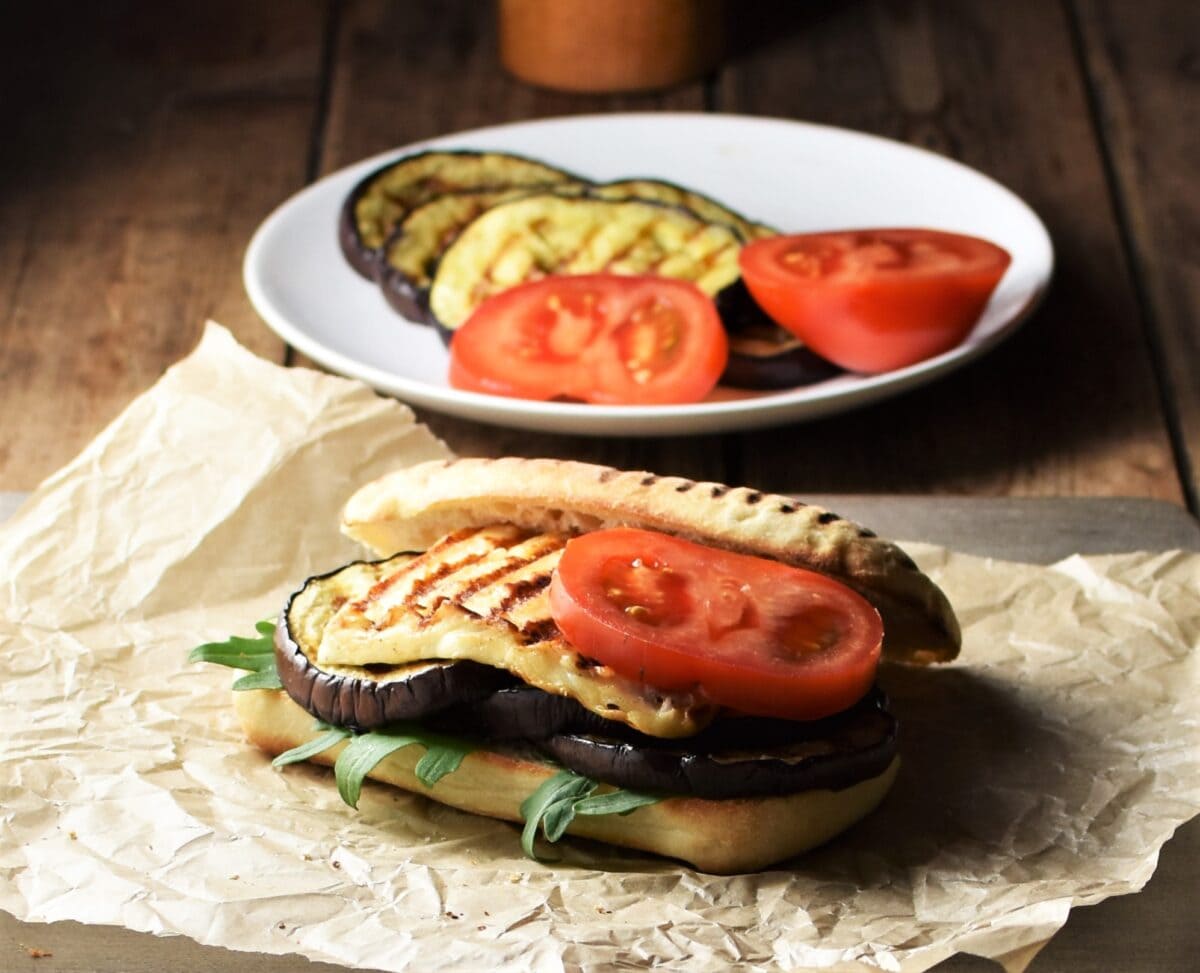 Side view of open eggplant halloumi tomato sandwich on top of paper, with grilled eggplant slices and tomato in background.