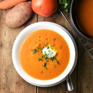 Creamy red vegetable soup in white bowl with sweet potato, carrot, tomato and pot in background.