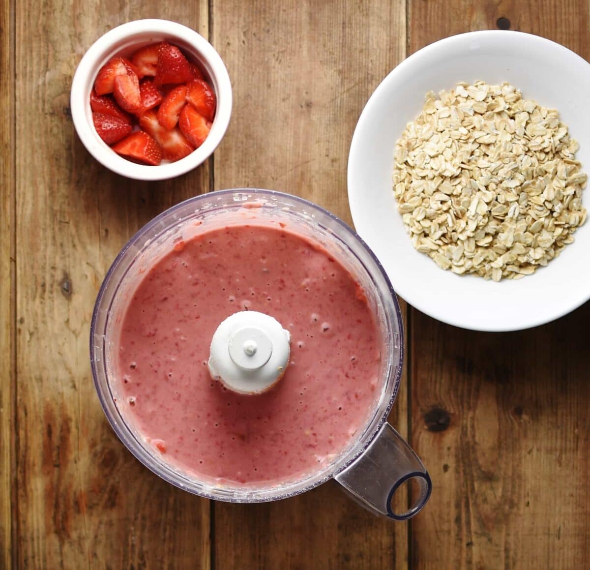 Top down view of pureed strawberry mixture in blender with oats in white bowl and strawberries in white dish.