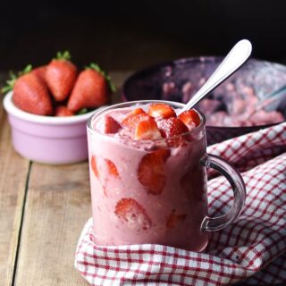 Side view of strawberry overnight oats in glass with spoon wrapped in red-and-white checkered cloth, with strawberries in pink dish and oats mixture in background.