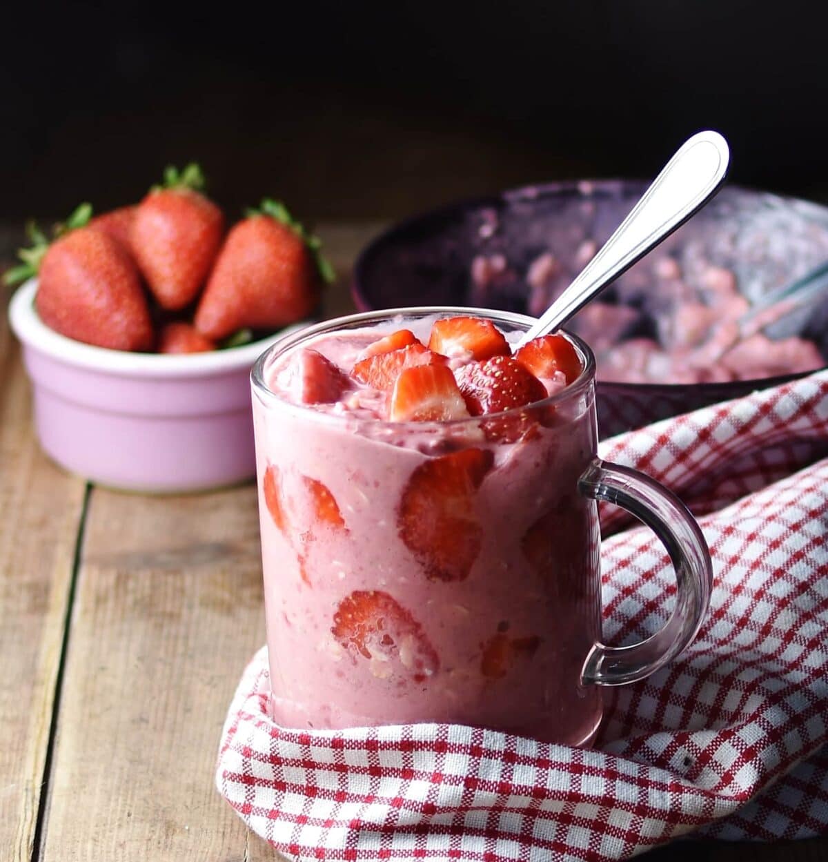 Side view of strawberry overnight oats in coffee cup with spoon wrapped in red-and-white checkered cloth, with strawberries in pink dish and oats mixture in background.