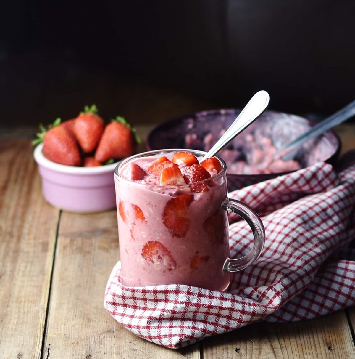 Side view of strawberry peanut butter overnight oats in coffee cup with spoon wrapped in red-and-white checkered cloth, with strawberries in pink dish and oats mixture in background.