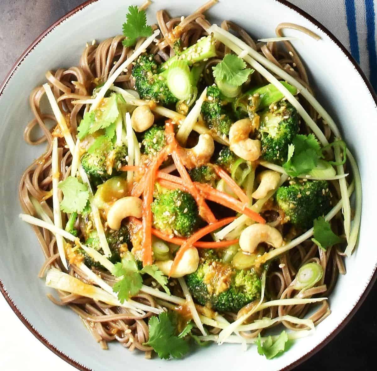 Top down view of broccoli and noodle salad with herbs, vegetables and nuts in blue bowl.