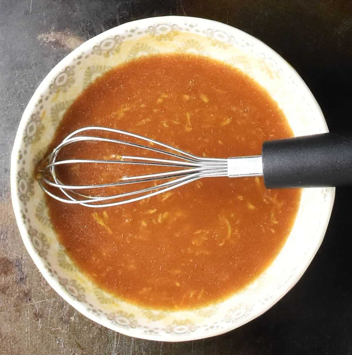 Brown coloured salad dressing in grey bowl with small whisk.