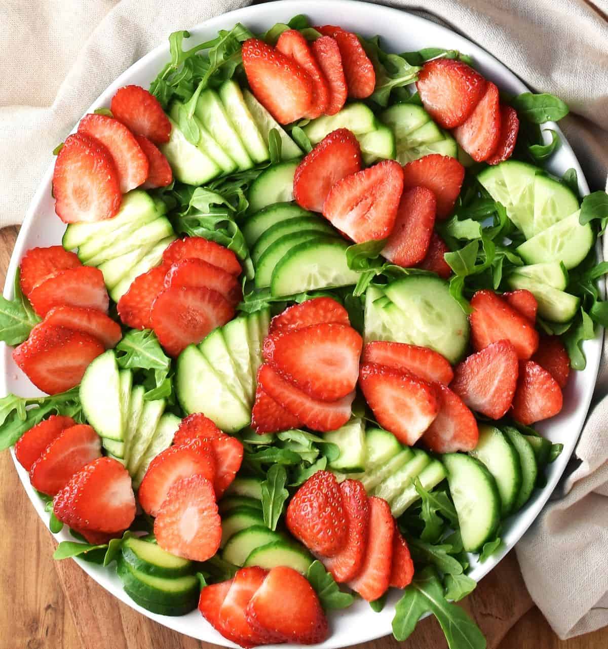 Top down view of sliced strawberries and cucumber with arugula on large white plate.