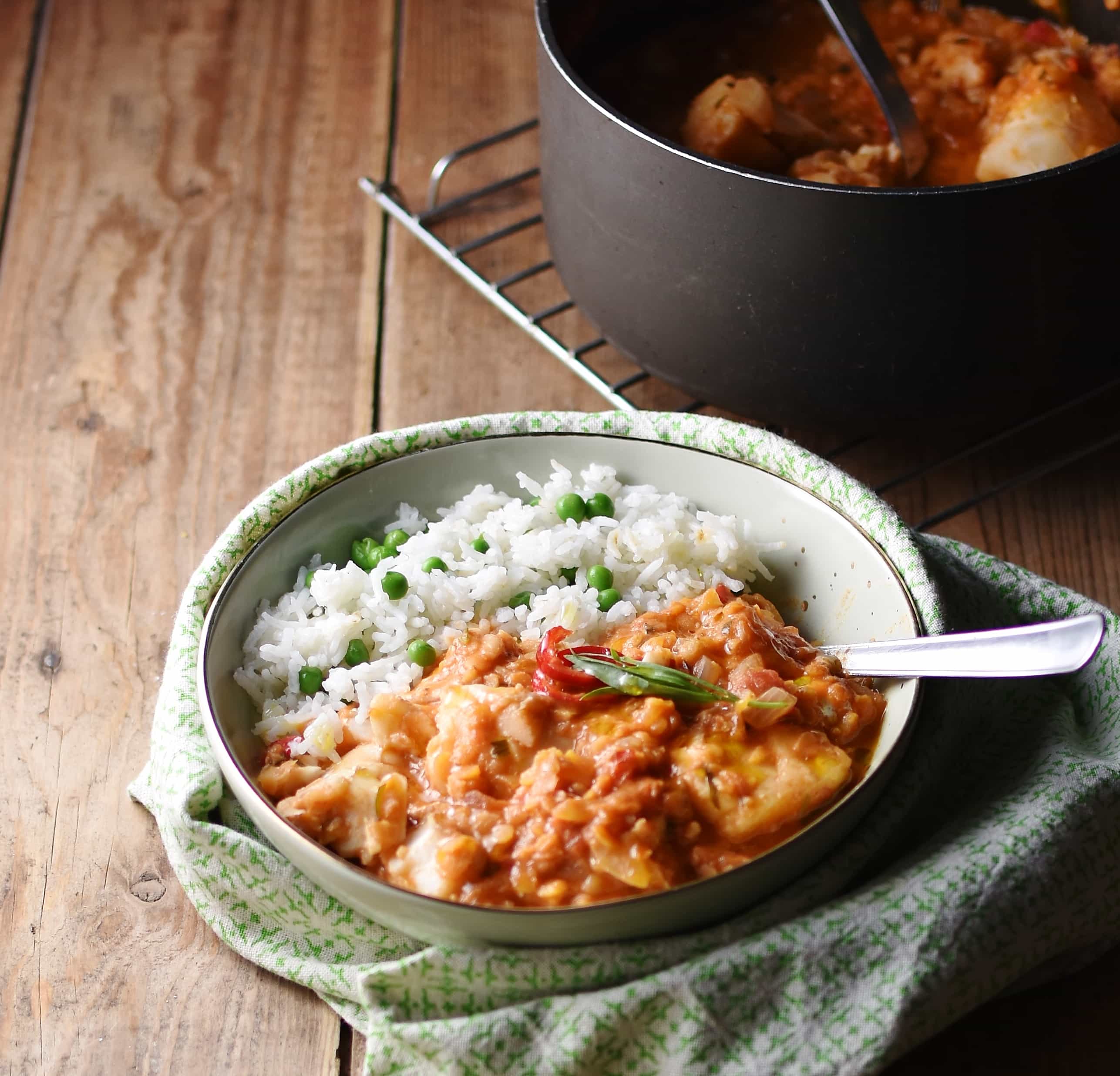Side view of tomato stew with rice and peas inside light green bowl with spoon wrapped in green cloth, with pot on top of cooling rack in background.