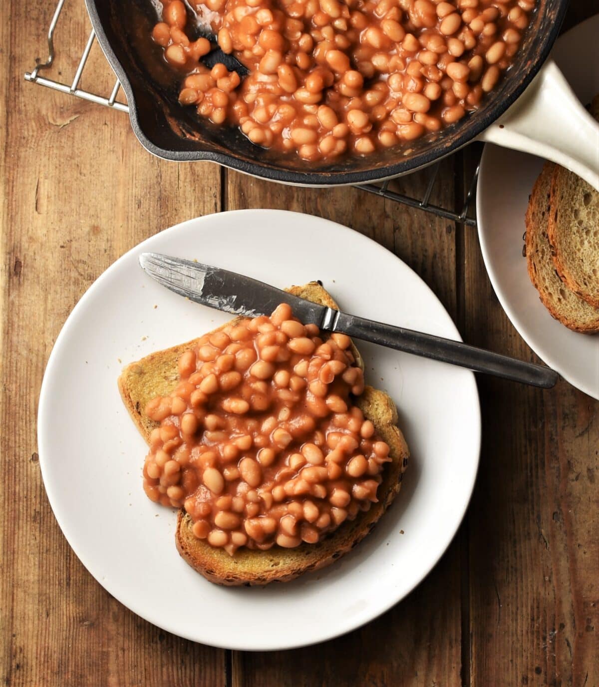 Baked beans on top of toast on white plate with knife, with beans in skillet in background.