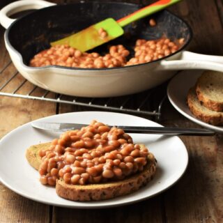Side view of toast with baked beans and knife on white plate with beans in skillet in background.