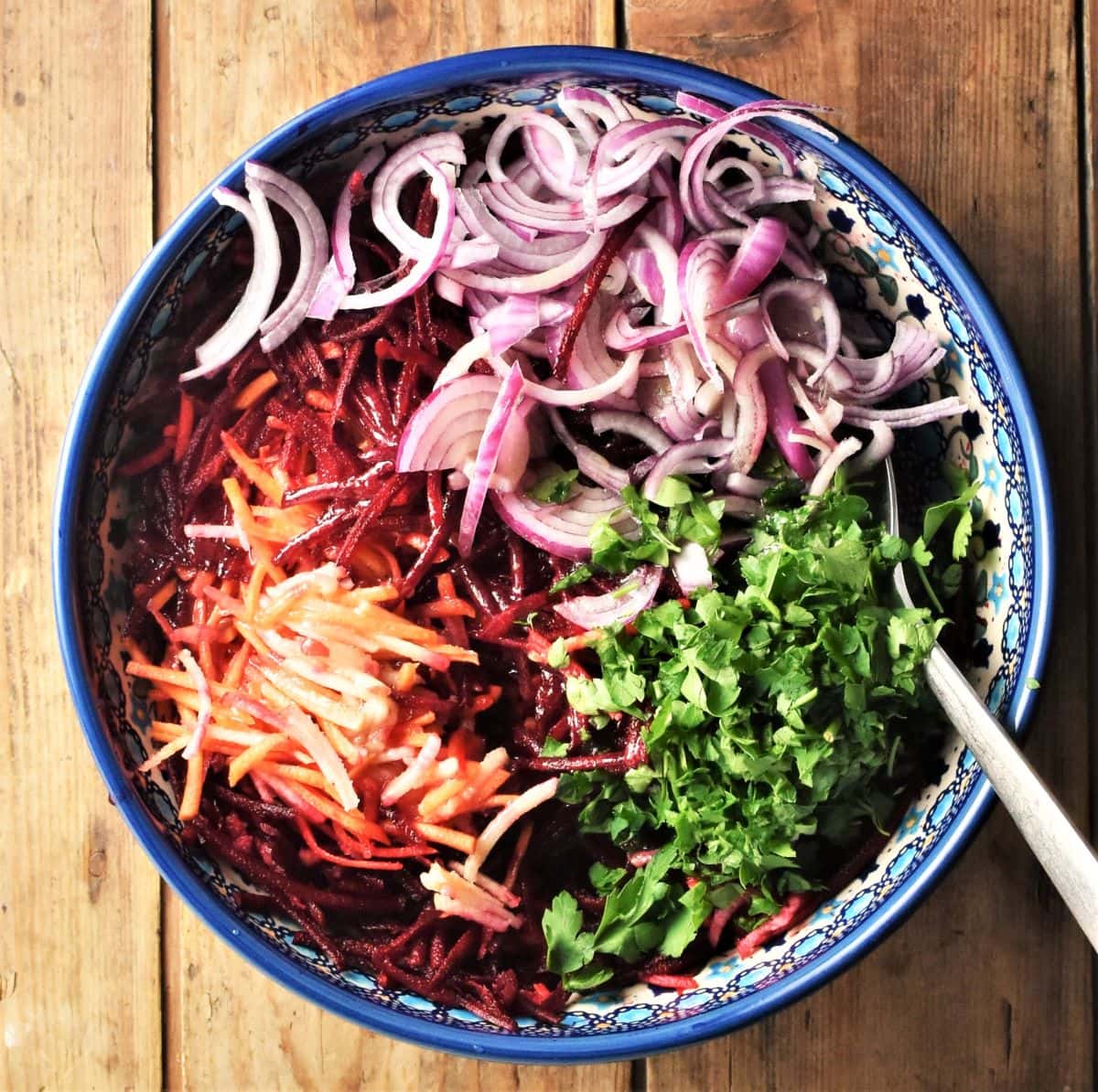 Shredded beetroot, sliced red onion and chopped parsley in blue bowl with spoon.