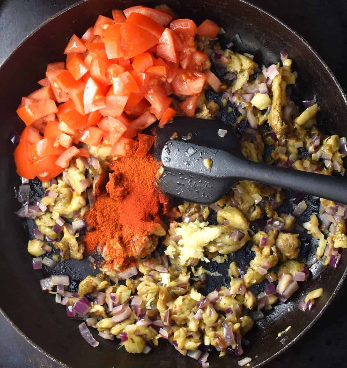 Cooking chopped eggplant with tomatoes and paprika in pan with black spoon.