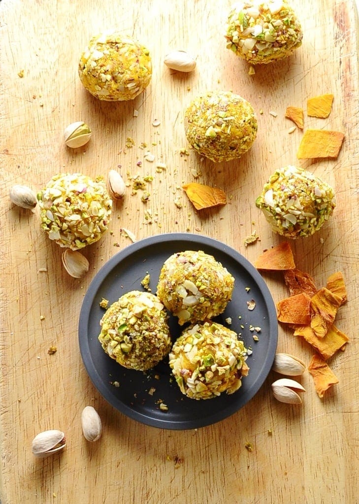 Top down view of mango cheesecake bites on small grey plate on top of wooden surface, with dried mango pieces and pistachios.