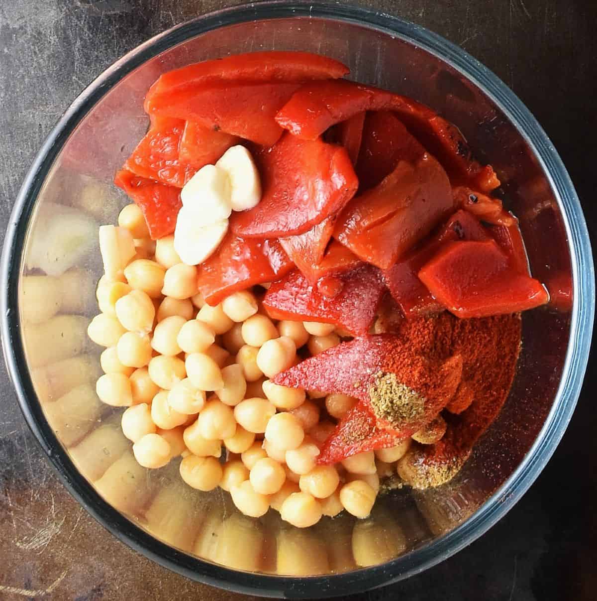 Chickpeas, chopped garlic and red roasted pepper in glass bowl.