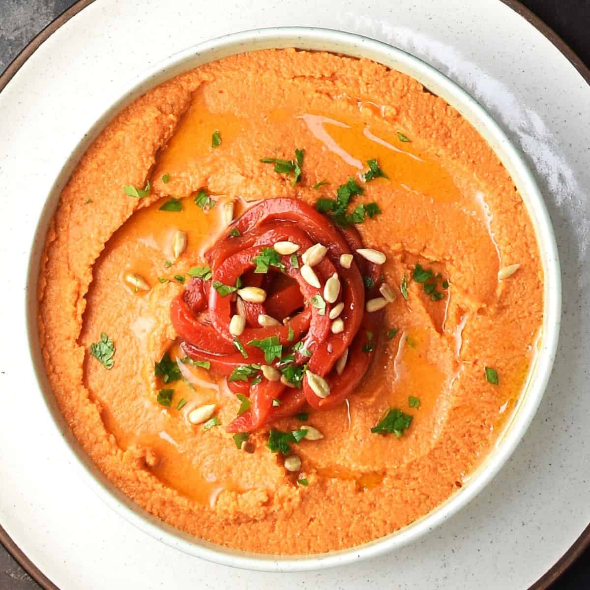 Top down view of roasted red pepper hummus without tahini garnished with peppers and seeds in white bowl.