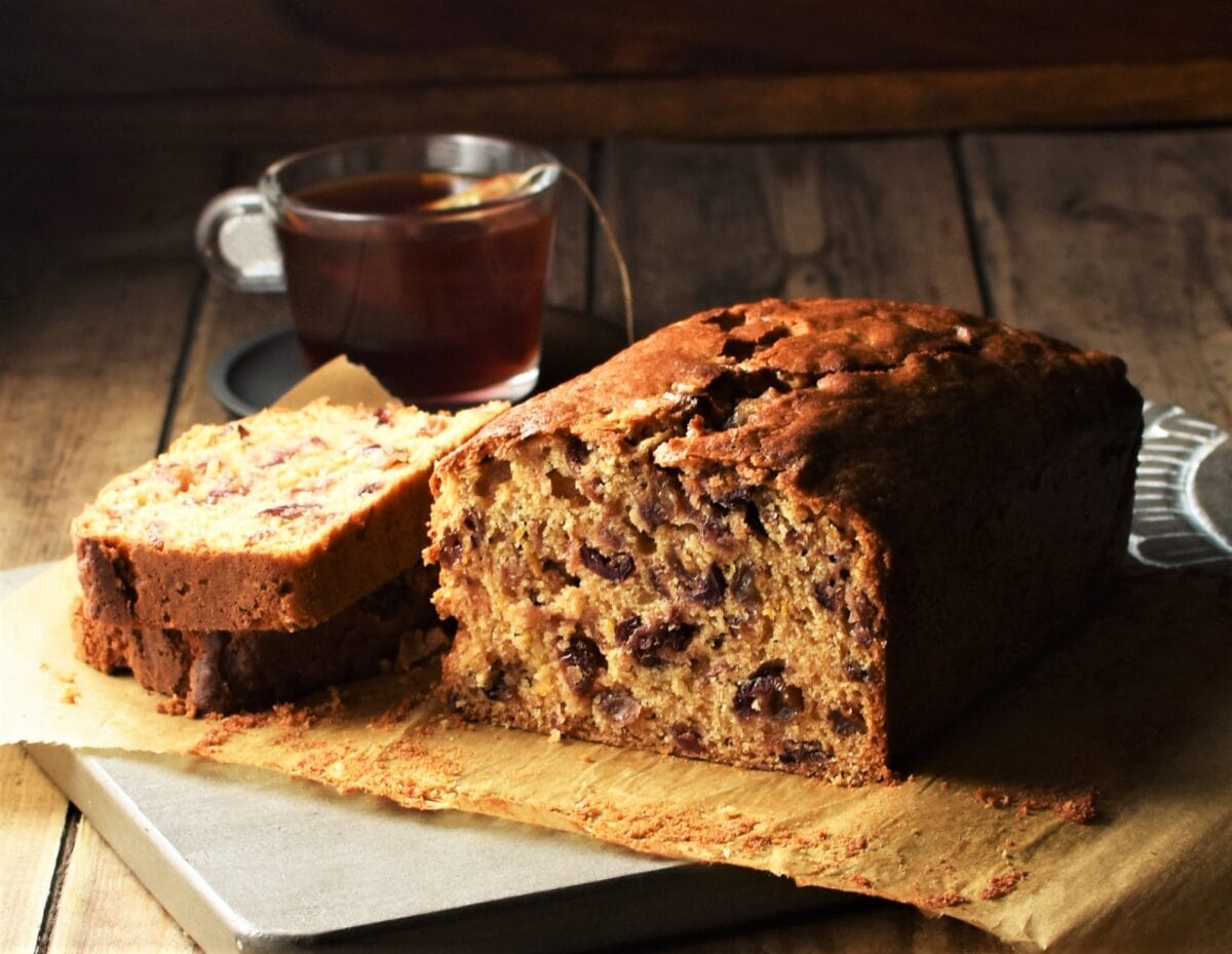 Side view of tea cake with slice on top of parchment, with cup of tea in background.
