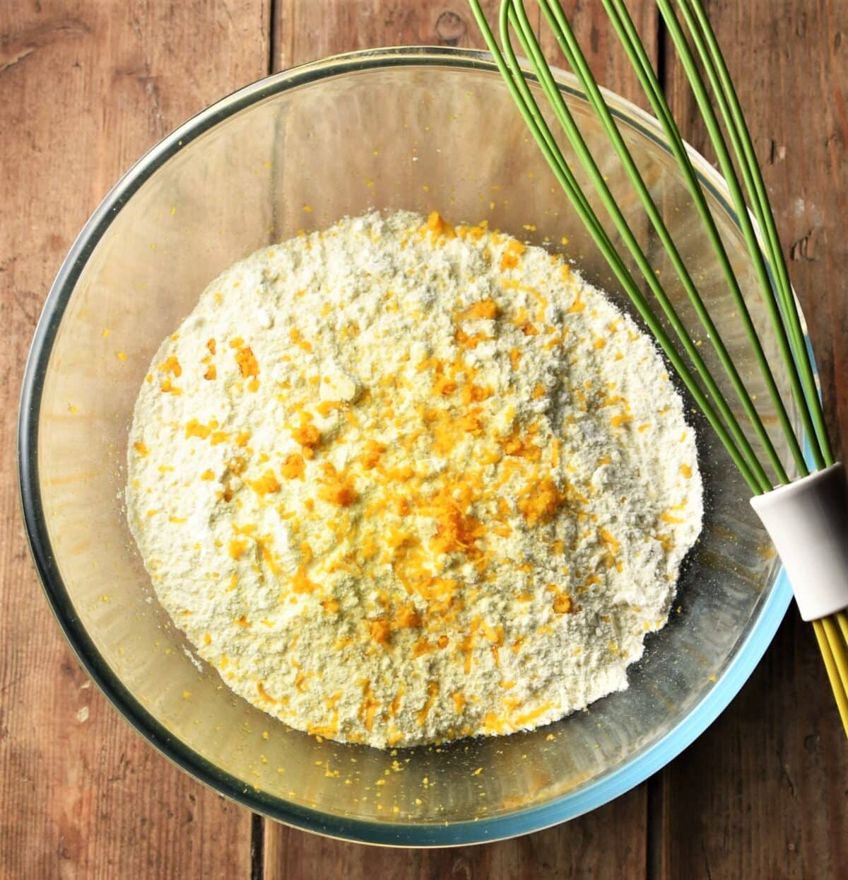 Flour mixture and orange zest in mixing bowl with green whisk on top.