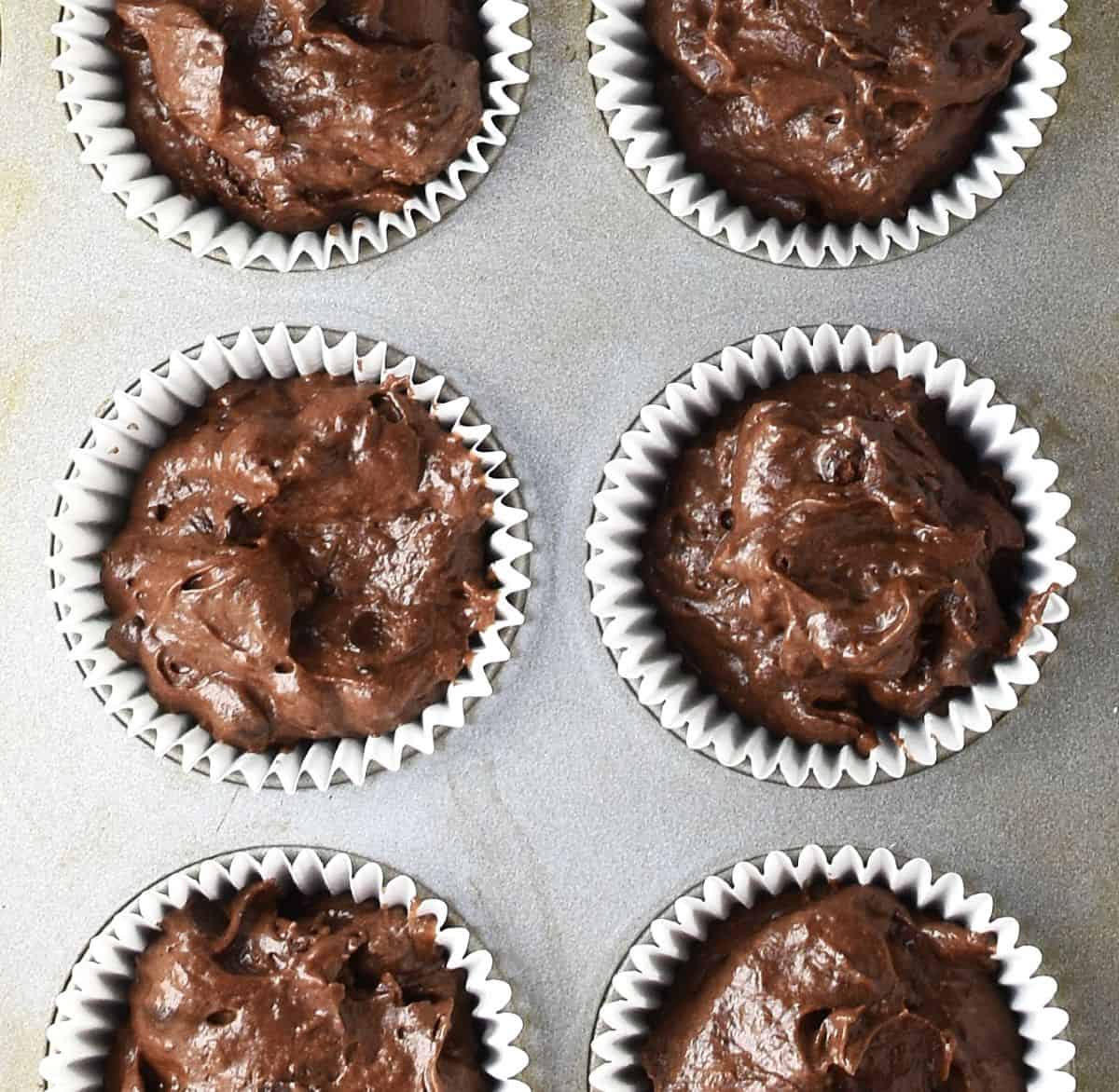 Top down view of chocolate avocado muffin batter in 6 white paper cases in pan.